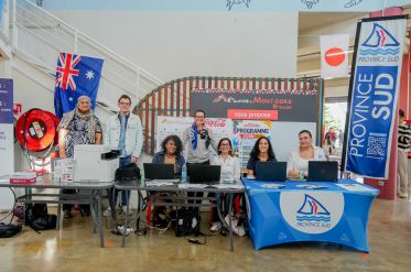 Group of six people standing and sitting behind a desk at an information booth, with banners reading Province Sud and Mont-Dore and various flags visible in the background (Australia flag on the left).