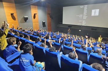 Large lecture hall filled with students in blue uniforms raising hands as a speaker presents at the front.