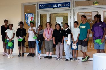 Group of teens and a woman posing outside a police station entrance, holding green caps. The door and a 'Accueil du Public' sign are visible behind them.